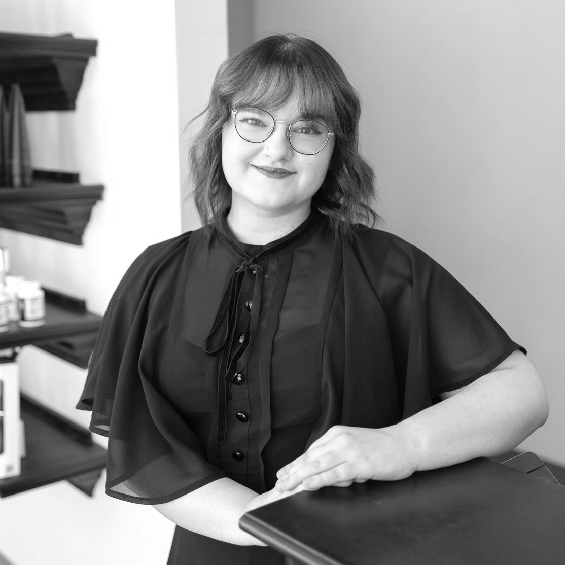 Black and White photo of a young woman short dark curls and bangs. She is wearing a pair of large, circular glasses, a dress with shawl sleeves, and a dark lipstick.