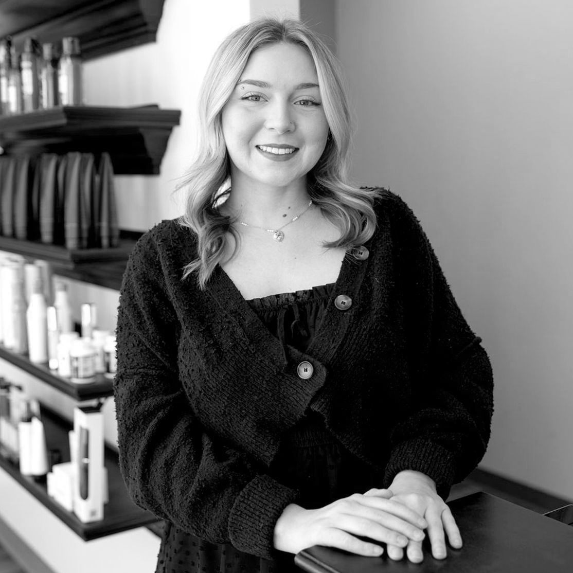 Black and White photo of a young woman with shoulder-length light-colored curls and a piercing on her right eyebrow. She is wearing a loose-fitting sweater buttoned over a chiffon dress.