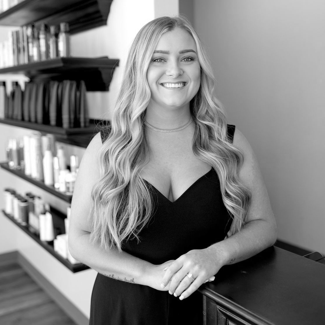 Black and White photo of a young woman with very long, light curls and a small tattoo of the moon on her forearm. She is wearing a simple dress with thick straps and a sweetheart neckline.
