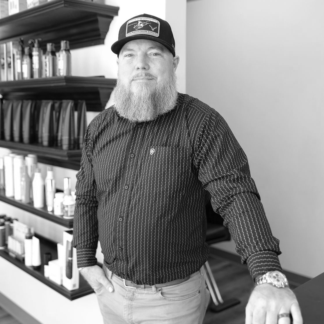 Black and White photo of a bearded man wearing a dark button-up with white stripes, a pair of khaki pants, and a baseball cap.