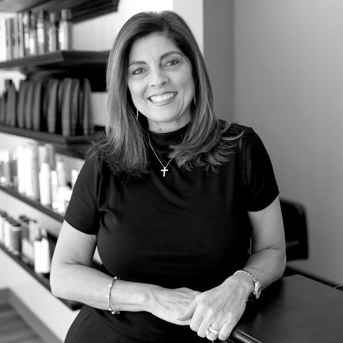 Black and White photo of an olive-complexioned woman with shoulder-length hair. She is wearing a short-sleeved mock turtleneck with a simple cross necklace.