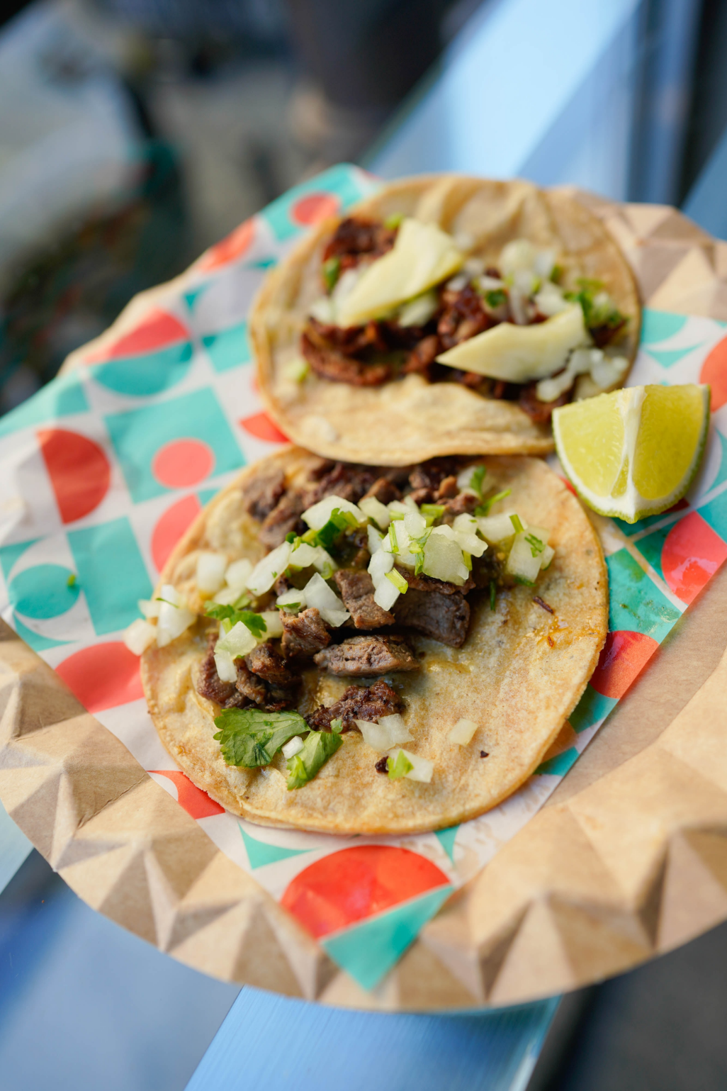 Two street tacos with fresh greens and plated with a lime on colorful paper.